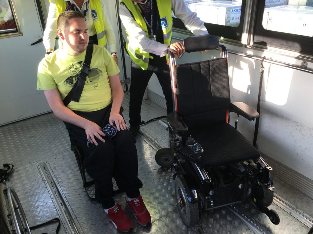 Rudy dans un fauteuil de transfert à côté de son propre fauteuil, à l'aéroport d'Athènes en 2016